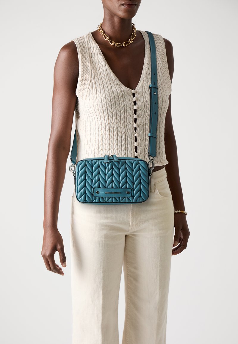 Geantă crossbody matlasată de culoare teal, cu formă rectangulară, accente din metal și curea ajustabilă, purtată peste un top crem din tricot cu model de cablu.
