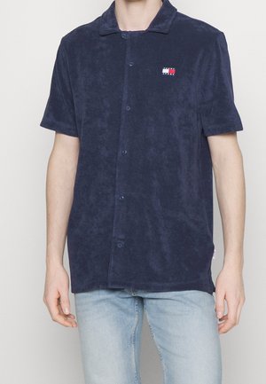 Muž nosící tmavě modrou krátkou košili na knoflíky s malým logem Tommy Jeans na levé straně hrudi a světlé modré džíny.