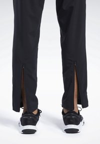 Černé sportovní kalhoty se zipovými otvory na kotnících, spárované s černobílými teniskami s texturovaným svrškem a zakřivenou podrážkou.