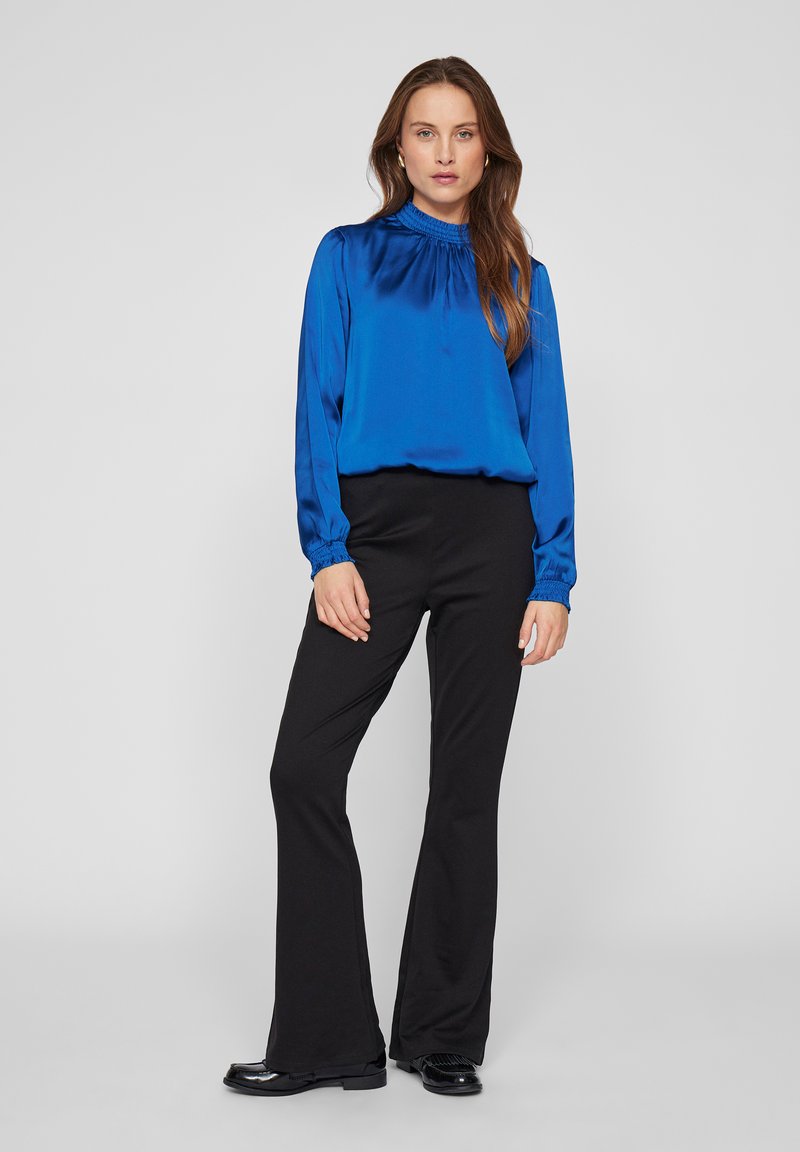 Vrouw die staat, gekleed in een blauwe blouse met lange mouwen en zwarte flare-broeken met zwarte schoenen, tegen een effen lichte achtergrond.