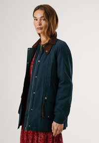Marineblauwe waterdichte jas met een bruine corduroy kraag. Heeft een ritssluiting aan de voorkant, twee zijzakken en een relaxte pasvorm. Gladde textuur.
