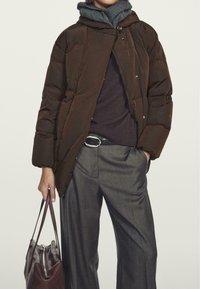 Brown puffer jacket with a hood, silver snap buttons, and a quilted texture, paired with a dark sweater and gray wide-leg trousers.