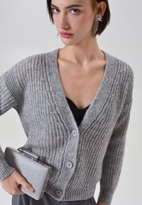 Cardigan grigio a maglia con scollo a V, cinque bottoni grigi, texture a coste e maniche lunghe. Il modello tiene in mano una borsa a tracolla argentata.