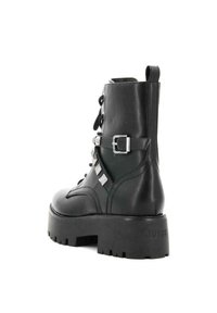 Bota de tobillo de cuero negro con cierre de cordones y hebilla, con tachuelas de plata a lo largo del costado y una suela de goma gruesa. Logo de la marca en el talón.