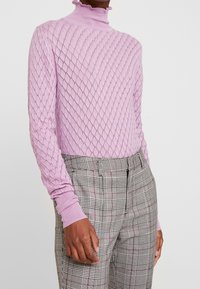 Personne portant un pull en maille à motif losanges violet clair et un pantalon à carreaux gris taille haute, debout devant un fond uni.