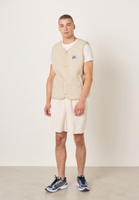 Smėlio spalvos flisinė liemenė su sagų užsegimu, dėvima ant balto marškinėlio. Derinama su šviesiais korduro kelnaitėmis ir mėlynais sportiniais batais.