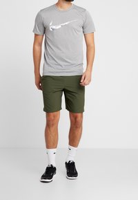 Camiseta gris de manga corta con un logo de Nike blanco y de camuflaje en el pecho, combinada con pantalones cortos verdes y zapatillas deportivas negras.