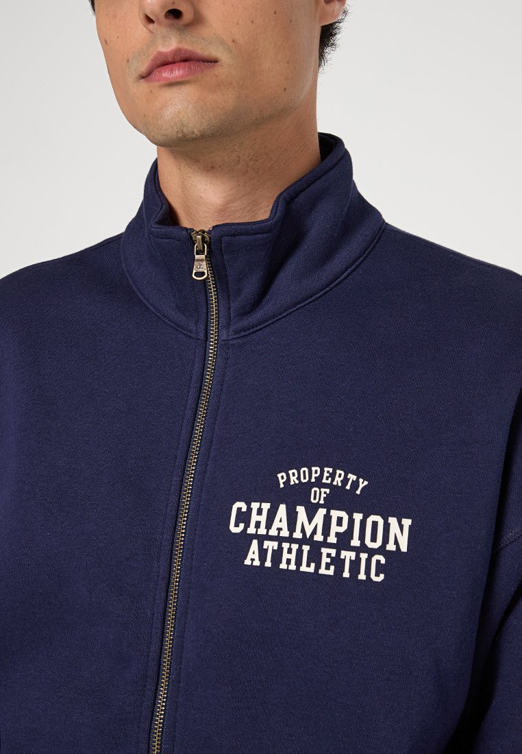 Uomo che indossa una felpa blu navy con zip con la scritta "Proprietà di Champion Athletic" stampata sul petto, mostrato dalla parte del collo al petto.