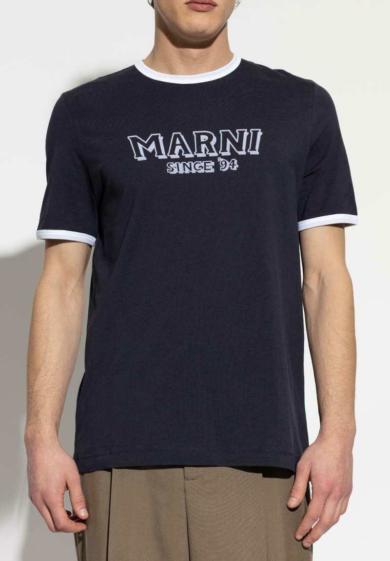 Homme portant un T-shirt noir à manches courtes avec des bordures blanches et le texte "MARNI SINCE '94" imprimé sur la poitrine, associé à un pantalon beige.