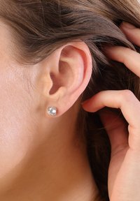 Boucles d'oreilles claires en cristal rond, en métal de couleur argentée, portées dans une oreille, avec une surface lisse et une finition polie.