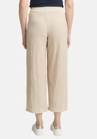 Pantalons larges beiges texturés à taille haute. Le matériau a un aspect froissé. Associés à des chaussures blanches avec des accents dorés.