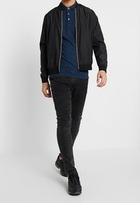 Svart bomberjacka med dragkedja, bärs över en mörkblå polo t-shirt, i kombination med figurnära svarta jeans och svarta träningsskor.