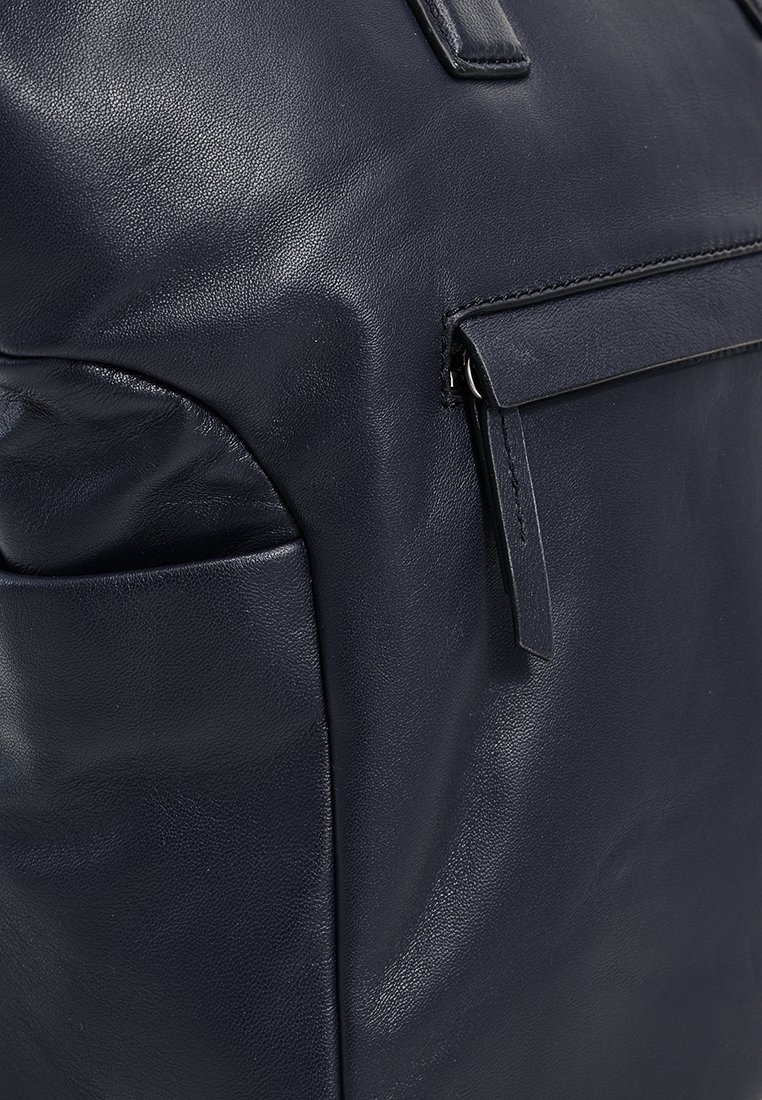 Zbliżenie na granatową skórzaną torbę, pokazujące przednią kieszeń na zamek błyskawiczny oraz boczną otwartą kieszeń z widocznym przeszyciem i teksturą.