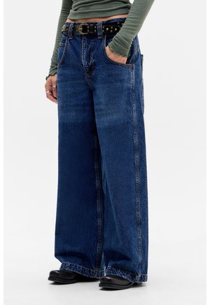 Person trägt weit geschnittene dunkelblaue Jeans mit einem schwarzen Nietengürtel, ein grünes, geripptes Langarmshirt, schwarze Schuhe und Ringe an der linken Hand.