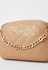 Bolso de mano acolchado beige con una correa de cadena dorada que presenta letras entrelazadas y un acento de cristal. Nombre de la marca "LIUJO" grabado en la parte frontal.