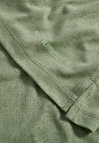 Hellgrüner strukturierter Stoff mit gefaltetem Saum und geprägt „hummel“-Logo nahe am Ärmelsaum.