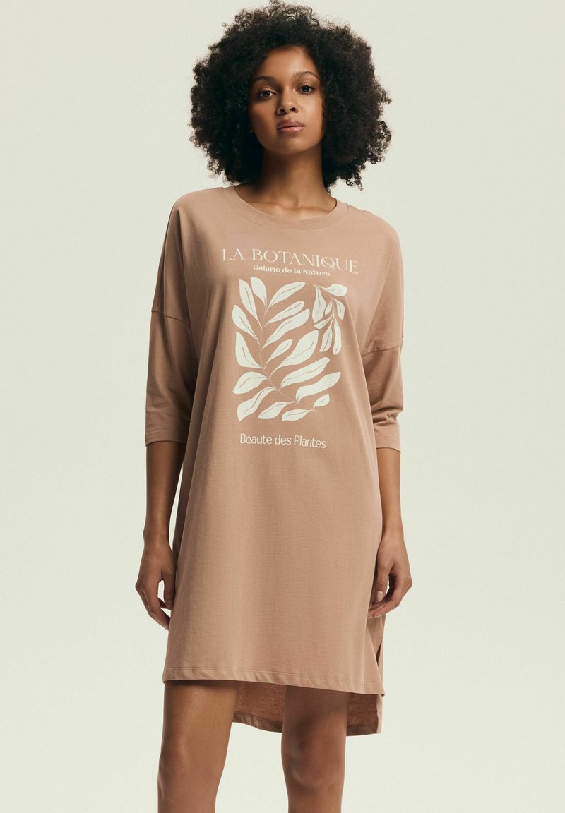 Kobieta stoi ubrana w luźną, beżową sukienkę o długim rękawie z roślinnym nadrukiem i francuskim tekstem z przodu, na tle jednolitego, jasnego tła.