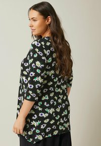 Blumen-Tunika mit schwarzem Hintergrund und weißen, lila und grünen Blumen, Dreiviertelärmel und lockerer Passform, aus weichem Stoff gefertigt.