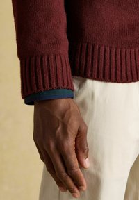 Ruka tmavej pokožky uvoľnená pri boku, nosiaca bordový pletený sveter cez vrstvený modrý manžet, proti krémovým nohaviciam a neutrálnemu pozadiu.