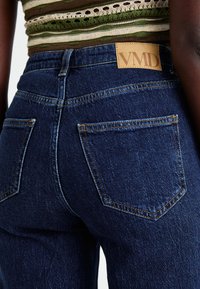 Dark blue denim jeans featuring a high waist, two back pockets, and a square leather tag with "VMD" stitched on the waistband.