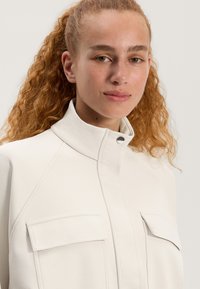 Weiße Jacke mit hohem Kragen, ausgestattet mit zwei Brusttaschen, einem strukturierten Stoff und einem klaren Design. Druckknopfverschluss-Detail am Kragen.