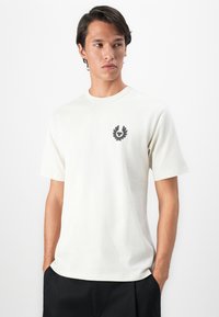 Ung mann iført en hvit kortermet t-skjorte med rund hals og et lite svart emblem på venstre bryst, kombinert med svarte bukser.