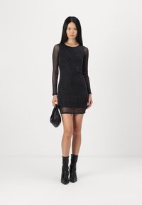 Μαύρο, μακρυμάνικο, πλεκτό φόρεμα με εφαρμοστή σιλουέτα, διαφάνειες στα μανίκια με υφή και όριο πάνω από το γόνατο. Συνδυασμένο με μια μαύρη τσάντα.