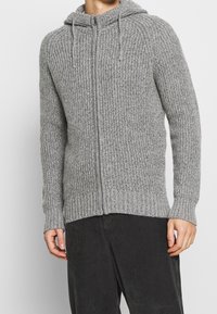 Grå stickad zip-up hoodie med ett texturerat ribbat mönster, en dragsko på huvan och åtsittande muddar, tillsammans med mörka kordbyxor.