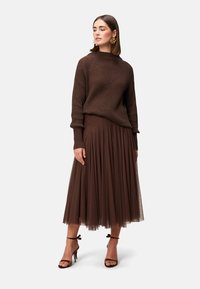 Maglione marrone a coste con sottili decorazioni, abbinato a una gonna in tulle marrone fluttuante. La modella indossa sandali alti marroni con cinturini.