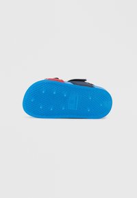 Niebieska gumowa podeszwa buta z teksturowanym bieżnikiem, o zaokrąglonym kształcie i małymi wypukłymi kropkami zapewniającymi przyczepność. Czarna i czerwona cholewka z paskami na rzepy.