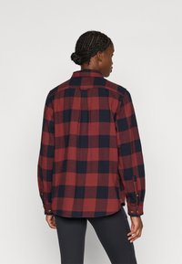 Camisa de flanela com estampa quadriculada em vermelho e azul-marinho, gola com botão, mangas compridas e punhos com botões visíveis; tecido texturizado com um ajuste descontraído.