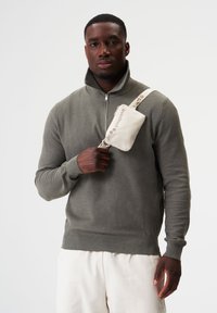 Olivgrüner Reißverschluss-Pullover mit strukturiertem Finish, kombiniert mit einer hellbeigen Umhängetasche mit Logo. Das Modell posiert in einer minimalistischen Umgebung.