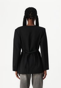 Černý blejzr s přiléhavým střihem, dlouhými rukávy a detailem na zavázání v pase. Hladký materiál se strukturovaným střihem. Šedé kalhoty doplňují vzhled.