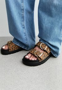 Sandaler med leopardtryck, två breda remmar, gyllene spännen och en tjock svart sula, bärs med blå vida jeans.