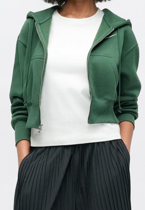 Persona che indossa una felpa verde con zip sopra una camicia bianca con pantaloni neri plissettati, mani in tasca, ritagliata al collo e in vita.