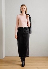 Žena u svijetlo ružičastoj majici dugih rukava, crnoj dugoj traper suknji visokog struka, crnim čizmama, drži crnu jaknu preko jednog ramena.