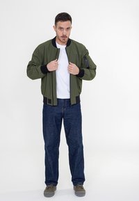 Oliwkowa bomberka z czarnymi wykończeniami, zapinana na zamek, z bocznymi kieszeniami, noszona na białej koszulce i niebieskich dżinsach. Szare buty.