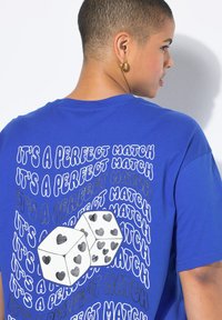 Modré bavlněné tričko s textem "TO JE PERFEKTNÍ SHODA" a grafikou dvou bílých kostek s černými srdíčky na texturovaném pozadí.