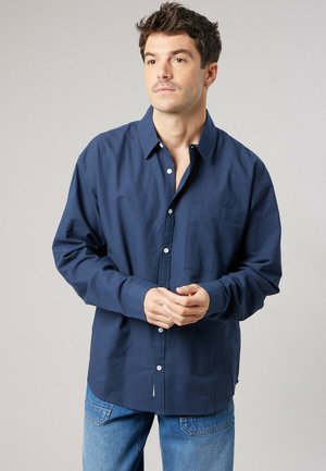 Man met kort donker haar, gekleed in een marineblauw overhemd met knopen en lichtblauwe jeans, staand met licht gevouwen handen.