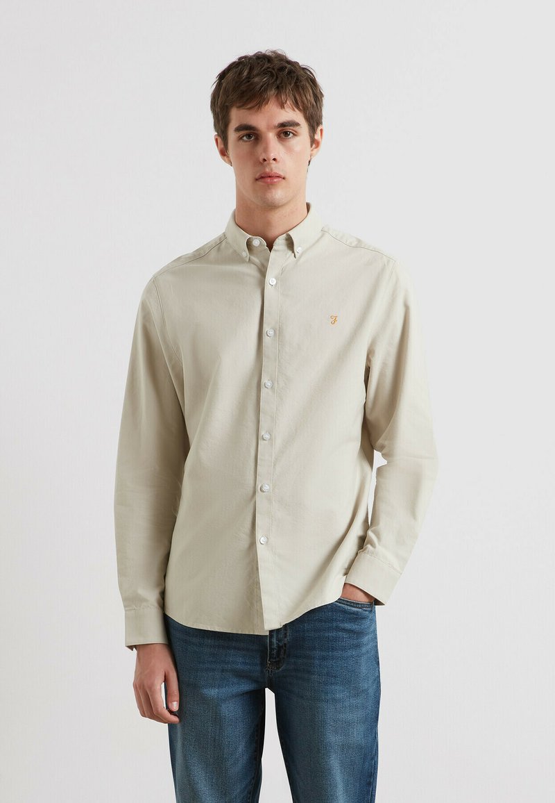 Beige overhemd met knopen en een kraag, voorzien van witte knopen en een klein geborduurd logo op de borst. G gedragen met blauwe spijkerbroek.