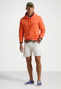 Oranž kapuutsiga dressipluus ees taskuga, valged teksapüksid, roheline nokamüts ja sinised spordijalatsid. Krae juures nähtav kihiline ruuduline särk.