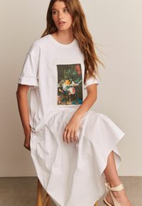Valge puuvillane t-särk, mis on dekoreeritud lillelise graafika printimisega, millel on sõna "Revival" kursiivis. Modell kannab valget voolavat seelikut ja heledat sandaali.
