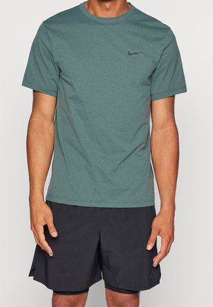 Mężczyzna ubrany w jednolity zielony t-shirt Nike i czarne sportowe spodenki, stojący z ramionami swobodnie opartymi wzdłuż ciała na jednolitym tle.