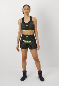 Black sports bra with neon green accents, paired with black shorts featuring a white and green Venum logo and geometric patterns, worn with black boots.