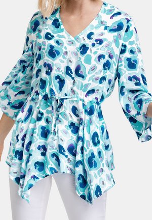 Kobieta ubrana w luźną tunikę z dekoltem w serek, z niebiesko-turkusowym abstrakcyjnym wzorem w panterkę, ze sznurkiem w talii i rękawami o długości do łokcia w kształcie dzwonka.