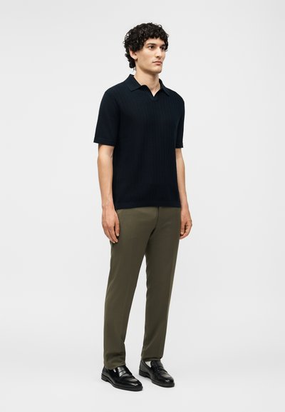 Άνδρας που στέκεται φορώντας μαύρο κοντομάνικο πόλο, ελαιδί παντελόνι και μαύρα επίσημα παπούτσια σε απλό ανοιχτό φόντο.