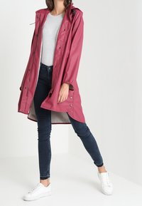 Różowy wodoodporny płaszcz przeciwdeszczowy z kapturem, zapięciem na napy i długim dołem, noszony na szarym t-shircie i ciemnych dżinsach, w zestawieniu z białymi sneakersami.