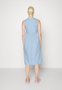 Vestido sem mangas de cor azul claro com um corpete ajustado e uma cintura franzida, atingindo a altura da metade da panturrilha. Tecido suave, design simples, usado com sapatos de lavanda.