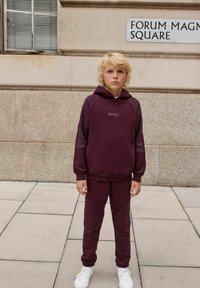Bordowa bluza z kapturem z logo, dopasowane spodnie i białe sneakersy; cechuje ją luźny krój i ściągacze u rękawów; plener w tle przy kamiennej ścianie.