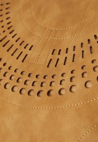 Lærmateriale i tan med perforerte sirkulære og lineære design. Glatt tekstur med kontrasterende sømmer. Subtil mønsterdetalj gjennom hele.
